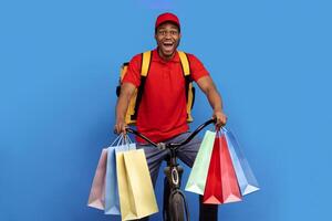 Delivery Service. Funny african american courier in red uniform and cap holding colorful shopping bags with customer's order while riding a bicycle with wide open mouth, blue studio background photo