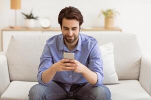 Man With Earbuds Using Phone Browsing Internet And Listening To Music Online Sitting On Couch At Home. Mobile User Texting On Smartphone In Living Room. Cellphone Application Concept photo