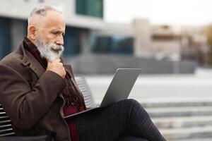 Pensive senior bearded businessman with laptop sitting on bench, typing on keyboard, sending email, empty space. Stylish aged grey-haired man sitting on street by business center, working online photo