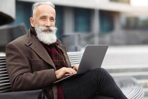 Wealthy grey-haired aged man with long beard in warm stylish outfit sitting on the bench by office building, using laptop, working online, chatting with clients or websurfing, empty space photo