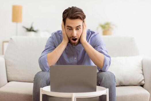 Shocked Businessman At Laptop Computer Touching Head Having Problem Working Online Sitting On Couch At Home. Distance Work Issue And Internet Connection Problems photo