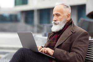 Concentrated grey-haired aged man with long beard in warm stylish outfit sitting on the bench by office building, using laptop, working online, chatting with clients or websurfing, empty space photo