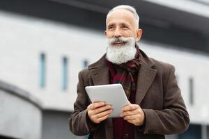 Cheerful aged man with long grey beard holding digital tablet while walking by street, looking at copy space and smiling. Senior businessman in stylish outfit with pad standing by office building photo