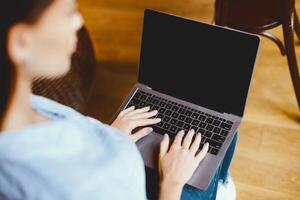 People And Technology Concept. Over the shoulder view of woman sitting and using laptop with black empty screen for mockup template, typing on keyboard, studying or working online at home photo