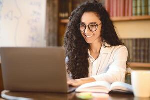 Freelance Concept. Portrait of smiling woman in eyeglasses using and working on computer, typing on laptop keyboard. Young lady sitting at desk with open notebook and doing work project photo
