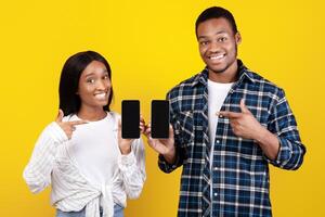 Online app for purchases and messages from stores. Smiling zoomers african american couple showing smartphones with blank screens and pointing fingers at gadgets, isolated on yellow background photo