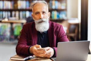 Wealthy elderly man using smartphone at cafe, sitting in front of laptop, freelancing or sending emails during lunch break. Aged businessman working online at cafe, using computer and mobile phone photo