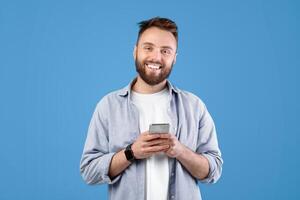 Portrait of cool bearded guy using mobile phone on blue studio background. Cheerful young man with modern gadget browsing internet, checking email, posting in social media photo
