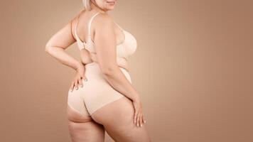 Young plus size woman in beige underwear posing over brown studio background, half-turned at camera, cropped image with free space photo