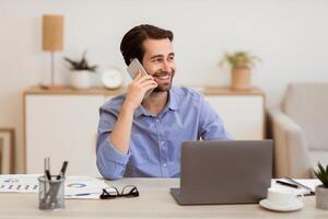 Cheerful Businessman Talking On Phone In Office, Sitting And Working On Laptop Computer. Happy Man Chatting On Cellphone At Workplace. Business Call, Mobile Communication Concept photo
