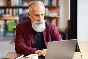 Aged businessman using laptop while having lunch break at cafe, drinking coffee, copy space. Elderly bearded man in stylish suit typing on laptop keyboard, working online or websurfing photo