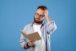 Young Caucasian student having difficulty understanding textbook, studying for exam on blue studio background. Puzzled millennial guy scratching head while reading complicated book photo