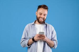 Portrait of positive Caucasian young guy using smartphone on blue studio background. Smiling millennial man with modern gadget chatting online, browsing web, posting in social media photo