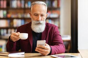 Grey-haired elderly man in burgundy jacket drinking coffee and using mobile phone, checking his email or chatting with clients, resting at cafe alone, empty space. Senior people and gadgets concept photo