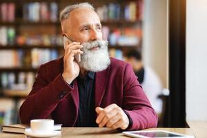 Busy aged businessman talking on mobile phone while drinking coffee at cafe, looking at copy space. Elderly handsome bearded man in suit spending time in lounge or cafe, having digital tablet photo