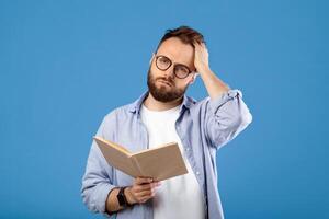 Studying is hard. Young bearded guy having problem understanding textbook on blue studio background. Millennial guy having headache while getting ready for exam or test photo