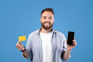 Positive millennial guy holding smartphone with empty screen and credit card on blue background, space for mobile app design. Young man promoting online shopping and contactless payment photo