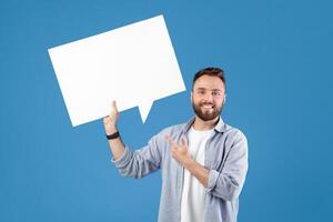 Happy young guy pointing at empty speech bubble with space for your ad or promo over blue studio background, mockup. Handsome Caucasian man with blank word cloud making announcement photo