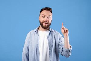Creativity, idea, inspiration. Young handsome guy pointing finger up, gesturing eureka, experiencing AHA moment on blue studio background. Cool bearded man finding solution to difficult problem photo