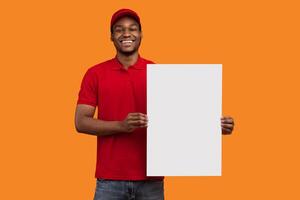 Shipment Service Promotion. Portrait of cheerful bearded black delivery man in red uniform holding and showing vertical empty blank board sign standing isolated on orange studio background photo