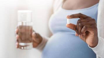 Supplements During Pregnancy. Unrecognizable Black Expectant Lady Holding Pill In Hand And Glass Of Water, Taking Maternity Vitamins For Health, Cropped Image With Selective Focus, Closeup photo