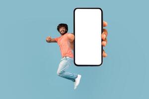 Happy handsome young eastern man jumping up and showing brand new cell phone with white blank screen over blue studio background, pointing at empty display, mockup, copy space photo