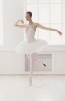 Classical Ballet dancer side view. Beautiful graceful ballerina practice ballet positions in tutu skirt near large window in white light hall. Ballet class training, high-key soft toning. photo