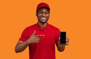 Choose Our Delivery Service. Smiling african american courier holding mobile phone with blank screen for mockup, wearing red cap and uniform, pointing at black empty gadget, orange studio background photo