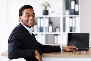 Successful african american businessman posing in his office, using modern laptop with blank screen, smiling at camera, copy space. Cheerful black manager in suit working on computer, mockup photo