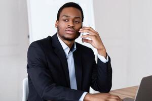 Pensive african american young entrepreneur having phone conversation with client or business partner in office, sitting at workdest with modern laptop, having bad news about his business, copy space photo