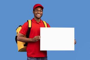 Portrait of cheerful bearded african american deliveryman wearing yellow thermo bag and red uniform holding and showing square empty blank board sign standing isolated on blue studio background photo