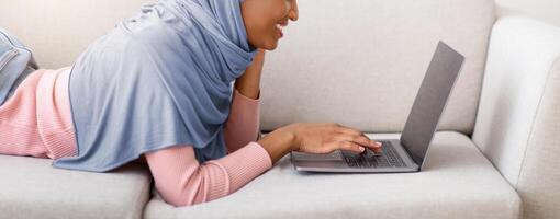 Side view of black woman in hijab using laptop with blank screen on sofa at home, copy space. African American lady in headscarf browsing internet, watching movie or communicating online photo