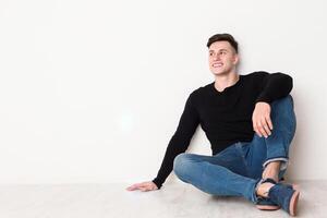 Smiling young man looks up, sitting on the floor at white background, copy space photo