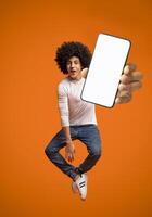 Funny young black teen guy jumping on air, showing cellphone with white empty screen on orange studio background. Young man demonstrating mockup copy space for your mobile app design photo