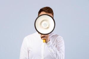 Faceless young businessman hiding behind megaphone on grey studio background. Millennial guy covering his face with loudspeaker. Public announcement or advertisement concept photo