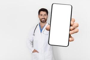 Medical App. Handsome Male Physician In Uniform Demonstrating Big Blank Smartphone At Camera, Smiling Doctor Man With Empty Cellphone In Hand Standing Over White Background, Mockup, Collage photo