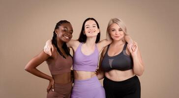 Group Of Body Positive Females With Different Figure Types Posing In Sportswear Over Brown Background In Studio, Three Happy Multi-Ethnic Women Embracing And Enjoying Their Beauty, Copy Space photo