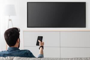 Young man watching television with blank screen and using smart TV remote control application on smartphone, back view. Select movie stream, change channel or settings in the menu photo