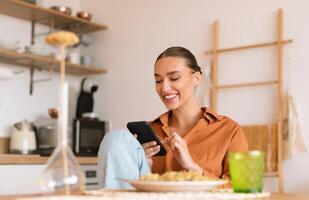 Excited lady using smartphone in kitchen while having dinner, sitting at table and texting on phone, browsing social networks or shopping online, free space photo