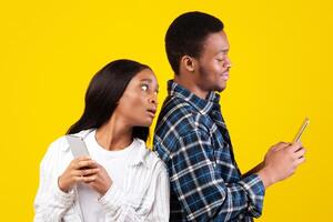 Jealousy, online networks and relationships. Interested millennial african american student lady in casual looks into phone of smiling husband, isolated on yellow background, empty space, studio shot photo