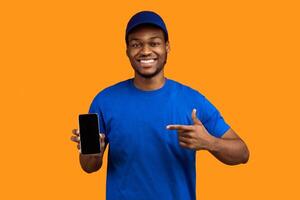 Recommending Service. Smiling african american delivery guy holding smartphone with blank screen for mockup, wearing blue cap and uniform, pointing at black empty monitor isolated on orange background photo