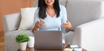 Online Briefing And Virtual Conference. Portrait of confident middle-aged woman sitting on the sofa, using laptop computer on desk, having call, talking to webcam, explaining and discussing photo