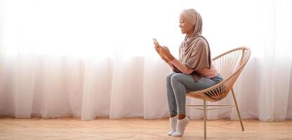 Millennial black lady in hijab using cellphone at home, working or studying remotely, free space. African American woman in Muslim headscarf checking email, sending message, having online call photo