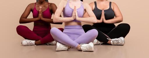 Group Of Three Women With Different Sizes And Skin Types Practicing Yoga Together, Beautiful Body Positive Females In Sportswear Meditating In Studio, Sitting In Lotus Pose Over Beige Background photo