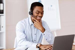 Positive young black man having online business conference in office, using laptop and headset, cheerfully talking to client or business partner, looking at computer screen and smiling, copy space photo