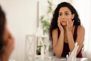 Beauty and skincare. Upset young woman looking in mirror and touching face, examining wrinkles and dark circles under her eyes at home, copy space. Dull tired skin concept photo