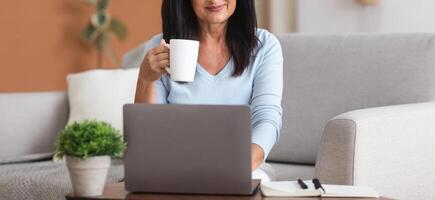Smiling middle-aged woman sitting on the couch, holding coffee mug and working on laptop. Businesswoman checking email. Beautiful lady and using computer while drinking a cup of tea photo