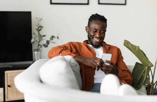 Cool gadget and app. Excited black man holding mobile phone and typing sms message, resting on sofa at home, free space. Man browsing internet and surfing web photo