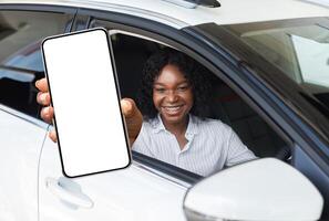 Car Renting App. Happy Black Female Showing Blank Smartphone While Sitting Inside Of Auto, Smiling African American Woman Demonstrating Copy Space For Mobile Advertisement Or Website, Mockup photo