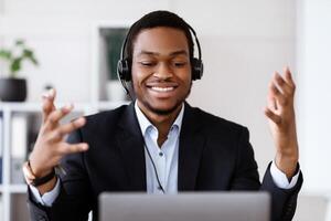 Closeup of inspired black businessman with headset having online conference, using laptop, talking to client or business partner, smiling at computer screen, business coach broadcasting from office photo